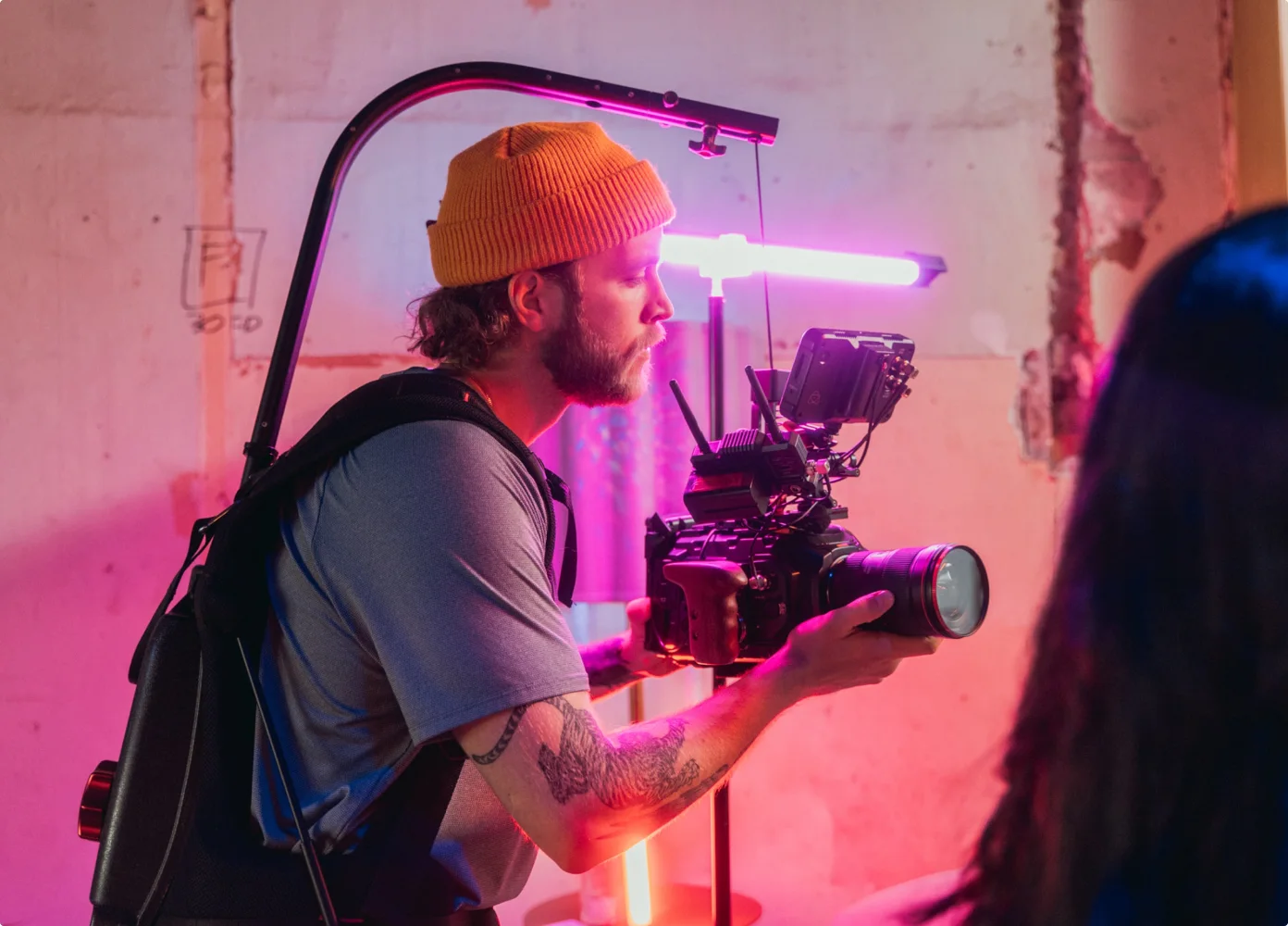 Ein Videofilmer mit gelber Strickmütze konzentriert sich intensiv auf eine hochwertige Kamera, umgeben von lebendigem rosa und violettem Licht