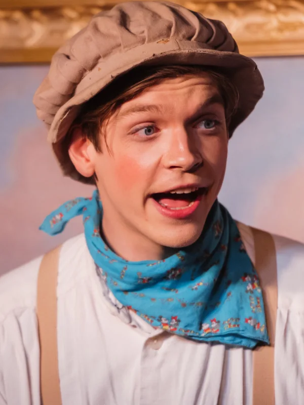 Ein junger Musicaldarsteller mit Hut und einem blauen Tuch um den Hals singt und spielt eine Rolle