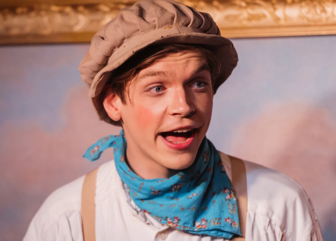 Ein junger Musicaldarsteller mit Hut und einem blauen Tuch um den Hals singt und spielt eine Rolle