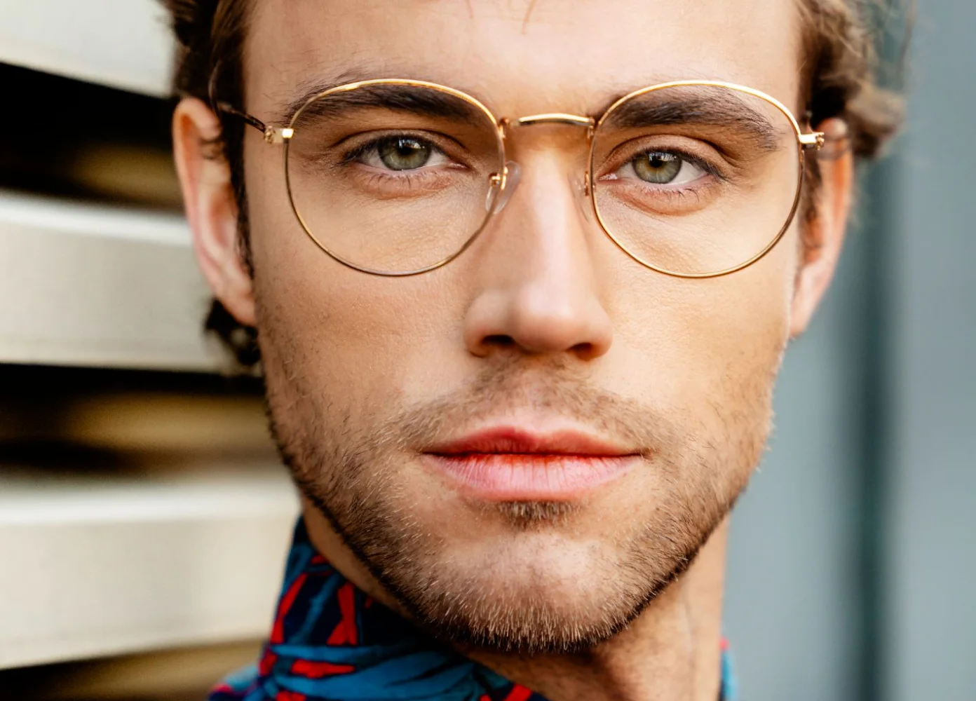 Ein männliches Model mit goldener Brille, grünen Augen und buntem Hemd schaut in die Kamera