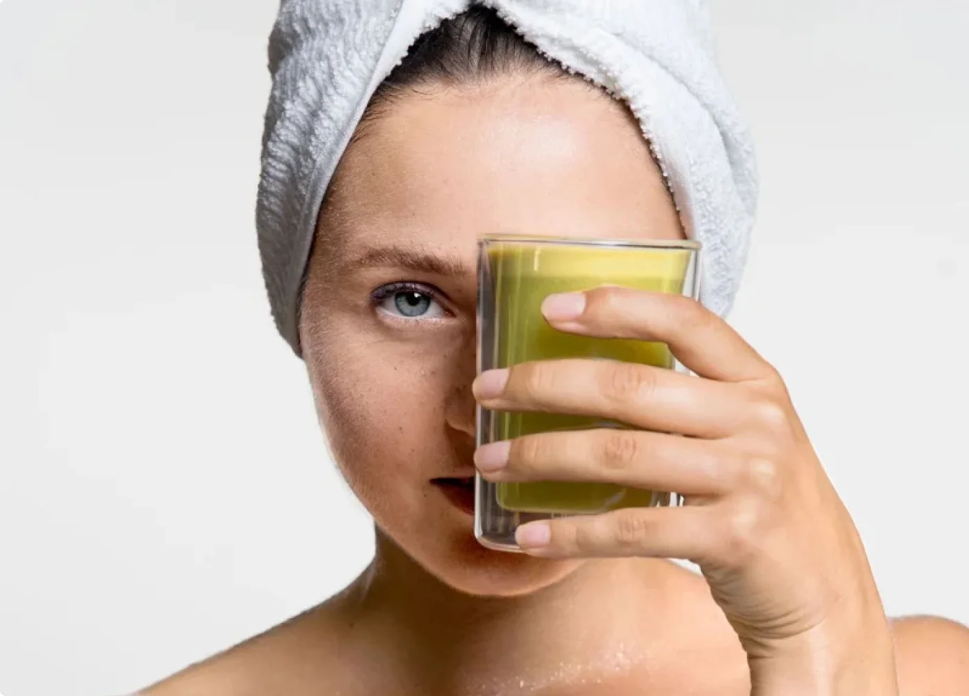 Eine weibliche Content Creatorin mit einem Handtuch auf dem Kopf hält ein Glas mit grünem Saft vor ihr Gesicht