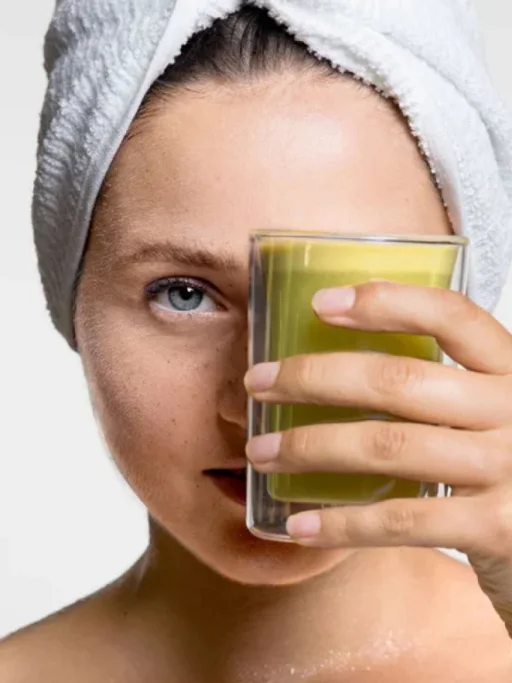 Eine weibliche Content Creatorin mit einem Handtuch auf dem Kopf hält ein Glas mit grünem Saft vor ihr Gesicht