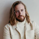 Mann mit Brille und langen Haaren trägt eine weiße Jacke und Rollkragenpullover