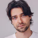 Mann mit mittellangem dunklem Haar und Bart trägt ein weißes T-Shirt und schaut in die Kamera