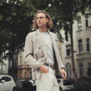 Mann mit langen Haaren trägt eine helle Jacke und Hose, steht auf einer Straße vor Wohnhäusern