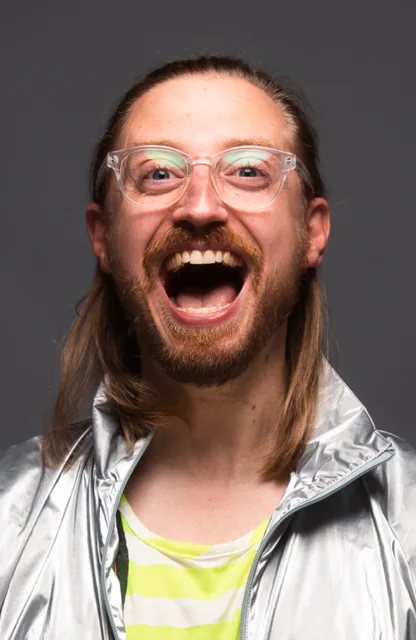 Ein Moderator mit Brille in einer silbernen Jacke und weit geöffneten Mund beim Lachen