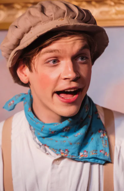 Ein junger Musicaldarsteller mit Hut und einem blauen Tuch um den Hals singt und spielt eine Rolle
