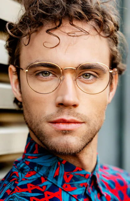 Ein männliches Model mit goldener Brille, grünen Augen und buntem Hemd schaut in die Kamera
