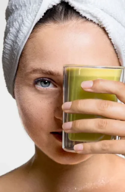 Eine weibliche Content Creatorin mit einem Handtuch auf dem Kopf hält ein Glas mit grünem Saft vor ihr Gesicht